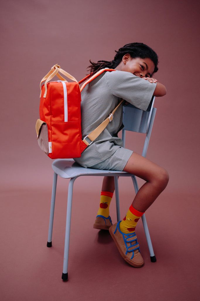 A child with dreadlocks sits backward on a chair, wearing a gray outfit, yellow and red socks, brown sandals, and carrying the Sticky Lemon Backpack Medium | Envelope | Campfire Orange—an orange recycled PET backpack with a waterproof lining—against a plain pink backdrop.