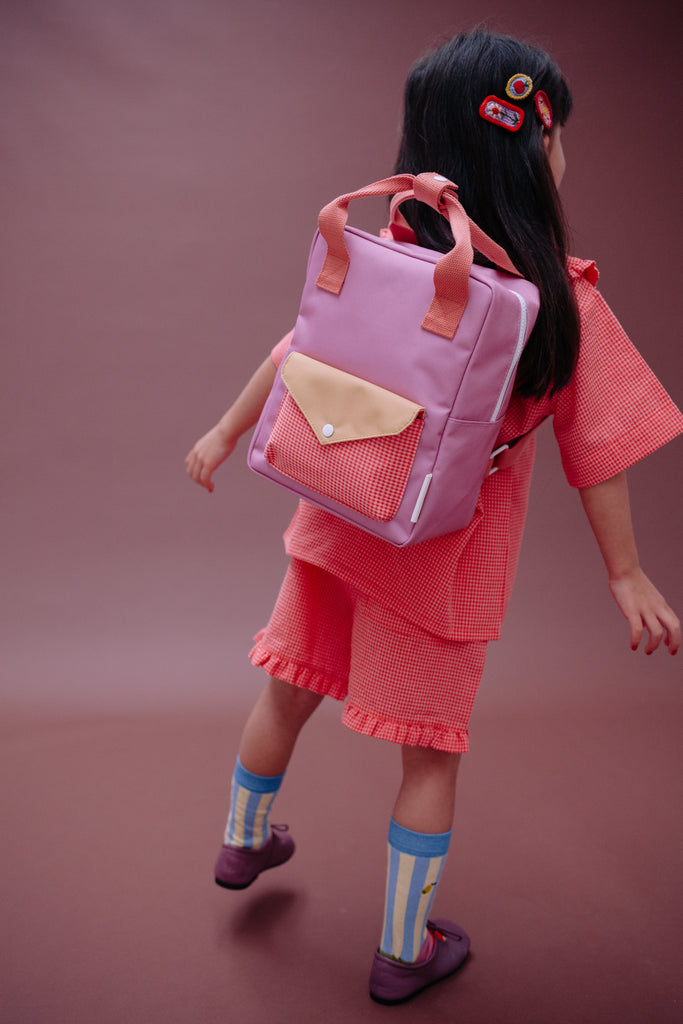 A young girl wearing a red outfit, colorful socks, and carrying the Sticky Lemon Backpack Small - Envelope - Rosebay Pink (ships in a week), featuring adjustable shoulder straps, walks away on a plain brown background.