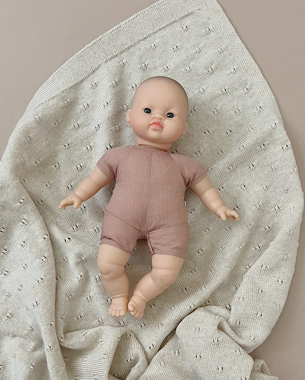 A Minikane Soft Body Doll (11") - Matteo with a bald head, wearing a light pink onesie, lays on a cream-colored, textured blanket with small perforations. Made from phthalate-free vinyl, this bedtime companion blends seamlessly into the neutral background.