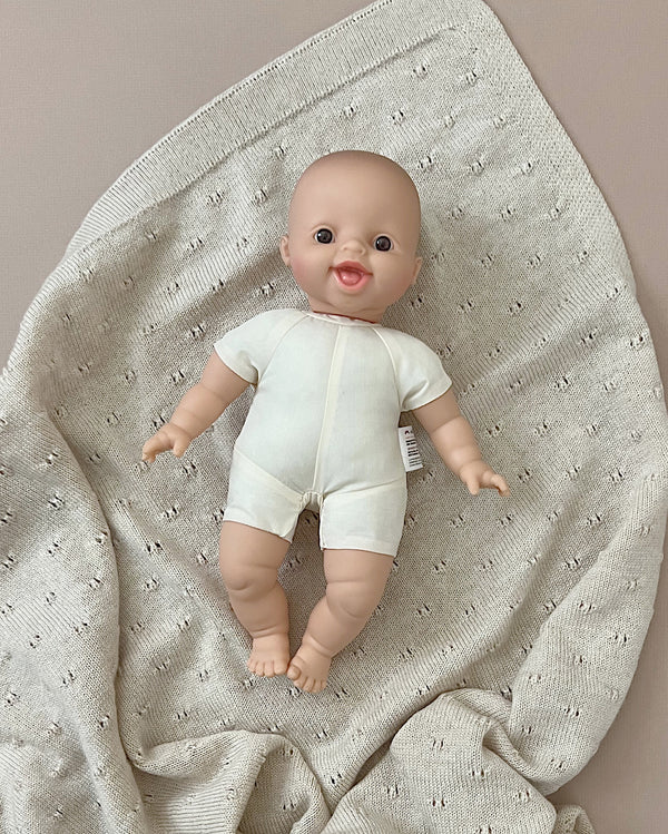 A Minikane Soft Body Doll (11") - Léo is lying on a textured beige blanket. The doll, perfect for imaginative playtime or as a bedtime companion, is wearing a simple white onesie and is positioned in the center of the knitted blanket, which has small holes in a repeating pattern.