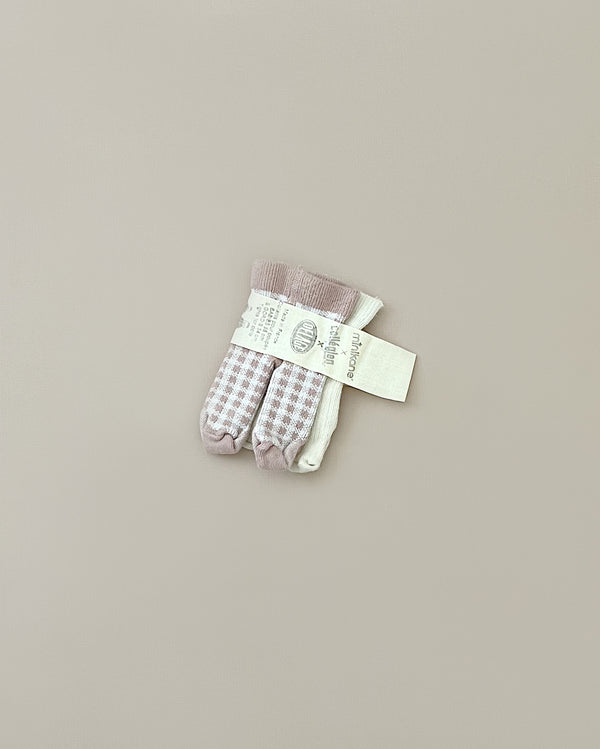A pair of Minikane Doll Clothing | Pack of Two Leggings in Ecru and Pink Checkers is neatly packaged on a plain beige background. The leggings have pink and white checkered patterns. A label with text is attached to the leggings, providing brand and product information, proudly stating they are French-made for a touch of elegance.