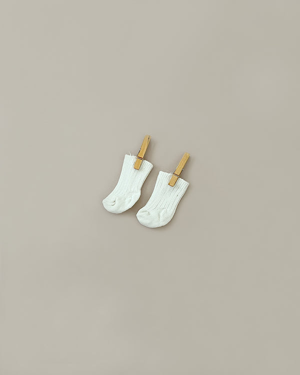 A pair of small white Minikane Doll Clothing | Beige Cotton Doll Socks is clipped to wooden clothespins against a neutral beige background. The socks, resembling Minikane Bambinis doll accessories, are suspended vertically by the clothespins, which create a minimalist and balanced composition.