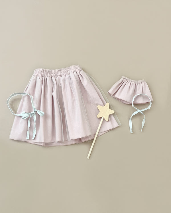 A Minikane | Little Fairy Costume In Petal Pink For Child and Doll is displayed on a beige background. The set includes an elastic-waist skirt, a matching smaller skirt, a headband with blue ribbons, and a wooden magic wand.