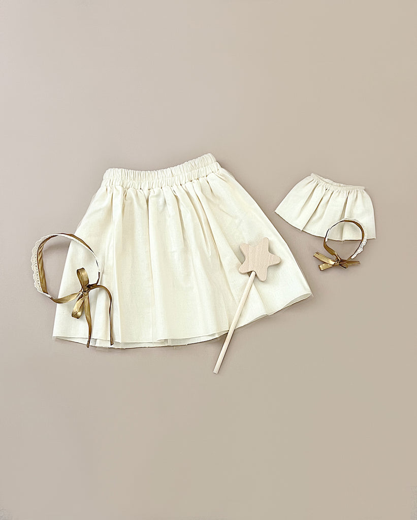 A flat lay photo featuring the Minikane | Little Fairy Costume In Linen Ecru For Child and Doll. Also included is a golden bow headband, a wooden toy wand with a star-shaped top, and charming fairy tutu elements perfect for any children's costume, set against a beige background.