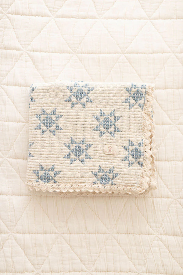 The Organic Cotton Baby Star Quilt - Sky, a cream-colored quilt with blue star patterns and scalloped trim, is displayed on a white, diamond-quilted surface. Ships in one week.