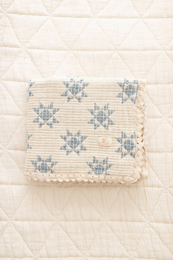 The Organic Cotton Baby Star Quilt - Sky, a cream-colored quilt with blue star patterns and scalloped trim, is displayed on a white, diamond-quilted surface. Ships in one week.