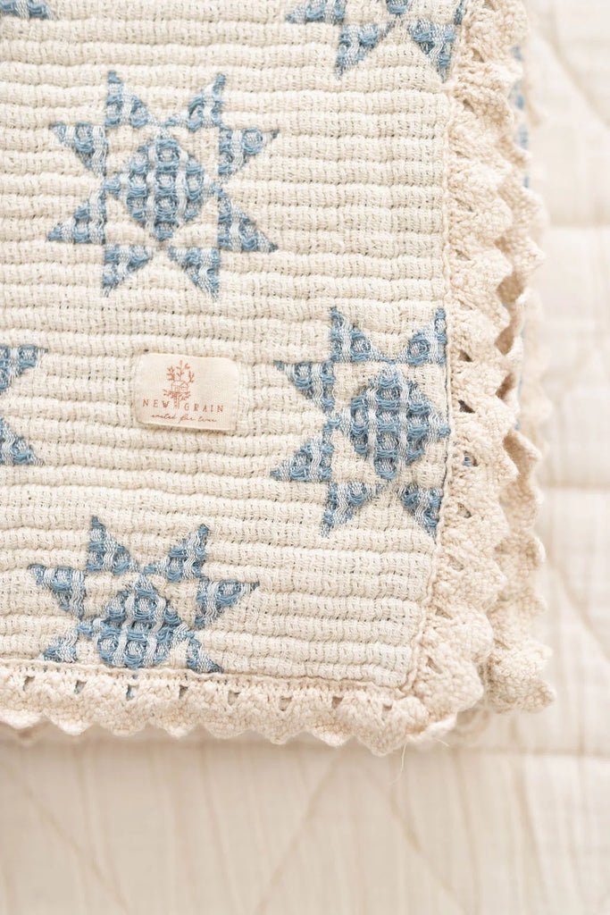 A close-up of the Organic Cotton Baby Star Quilt - Sky, featuring cream fabric with blue star patterns and delicate crochet trim. A small "NEW & BRAIN" tag is visible. The quilt is layered on a white, quilted surface. Ships in one week.