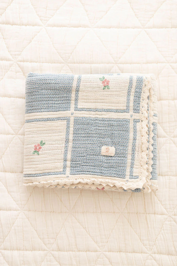 The Organic Cotton Patchwork Baby Blanket - Spring Bloom features a blue and white checkered pattern, small embroidered flowers, and crochet trim, shown folded on a white quilted surface.