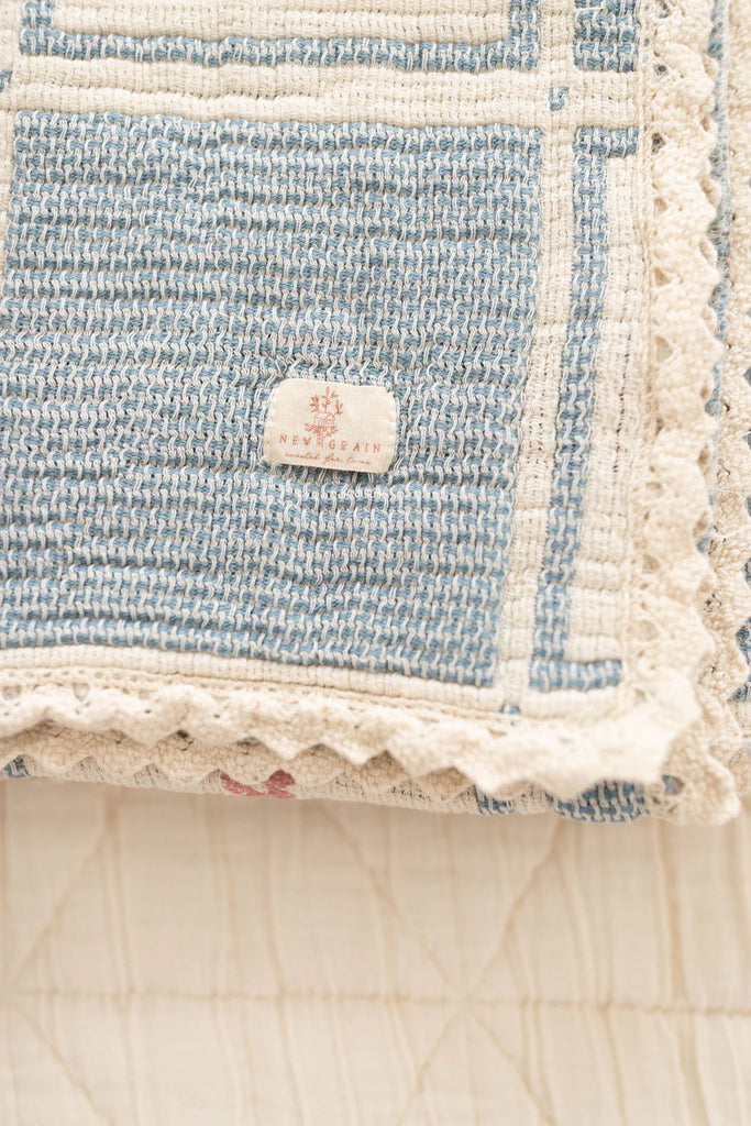A close-up of the Organic Cotton Patchwork Baby Blanket - Spring Bloom, showing its blue and cream crochet trim with a scalloped edge and a "REVIVAL" fabric tag on the corner, folded on a light quilted surface.