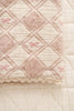 A close-up of the Organic Cotton Baby Patchwork Blanket - Pink Bows, featuring a diamond quilted pattern, pink and cream tones, embroidered bows, and scalloped edges, displayed on a matching textured surface. Ships in one week.
