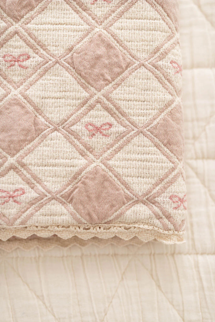 A close-up of the Organic Cotton Baby Patchwork Blanket - Pink Bows, featuring a diamond quilted pattern, pink and cream tones, embroidered bows, and scalloped edges, displayed on a matching textured surface. Ships in one week.