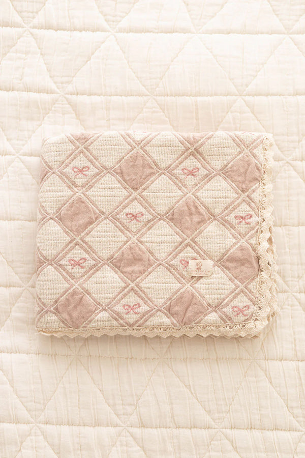 The Organic Cotton Baby Patchwork Blanket - Pink Bows (ships in one week), featuring a diamond quilted pattern and lace trim, is folded on a textured, cream-colored surface.