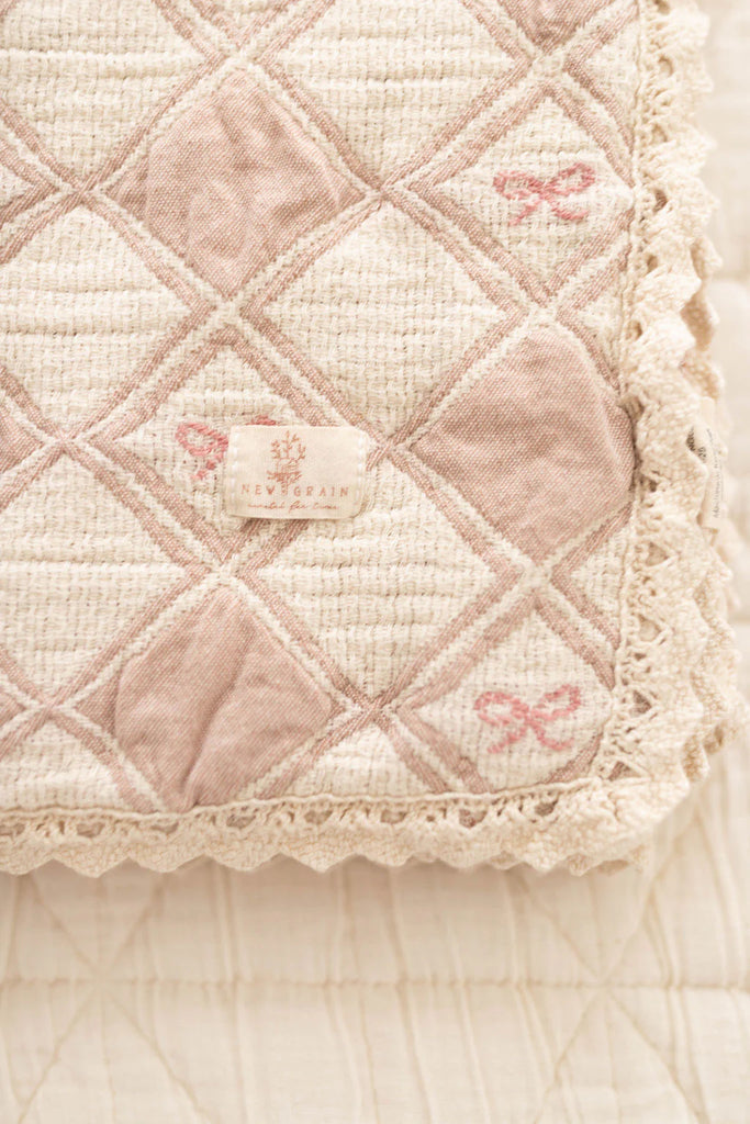 A close-up of the Organic Cotton Baby Patchwork Blanket - Pink Bows, featuring soft quilted cotton in beige and light pink with a diamond pattern, lace trim, and delicate details—perfect as a baby blanket. Ships in one week.