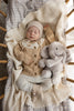 A baby in beige knitwear cuddles the Snuggle Bunnies - Olive The Elephant plush while resting on a cream blanket in a wooden crib. Gentle light from the side adds to the calm, cozy atmosphere.