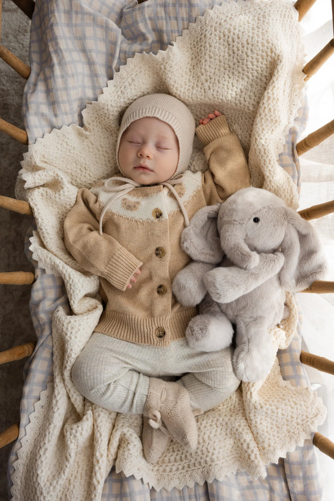 A baby in beige knitwear cuddles the Snuggle Bunnies - Olive The Elephant plush while resting on a cream blanket in a wooden crib. Gentle light from the side adds to the calm, cozy atmosphere.