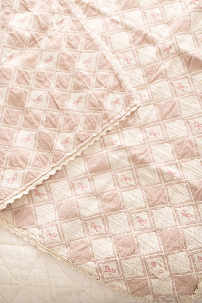 The Organic Cotton Baby Patchwork Blanket - Pink Bows features a soft pink and white design with embroidered diamond patterns and small bows, neatly draped on a bed. Ships in one week.