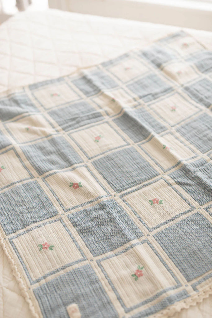 The Organic Cotton Patchwork Baby Blanket - Spring Bloom features soft, quilted organic cotton in blue and cream squares with embroidered pink flowers, displayed atop a lightly patterned bedspread.