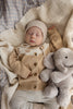 A baby in a beige knit hat and sweater sleeps on a cream blanket, cuddling Snuggle Bunnies - Olive The Elephant plush toy.