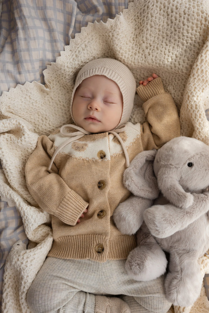A baby in a beige knit hat and sweater sleeps on a cream blanket, cuddling Snuggle Bunnies - Olive The Elephant plush toy.