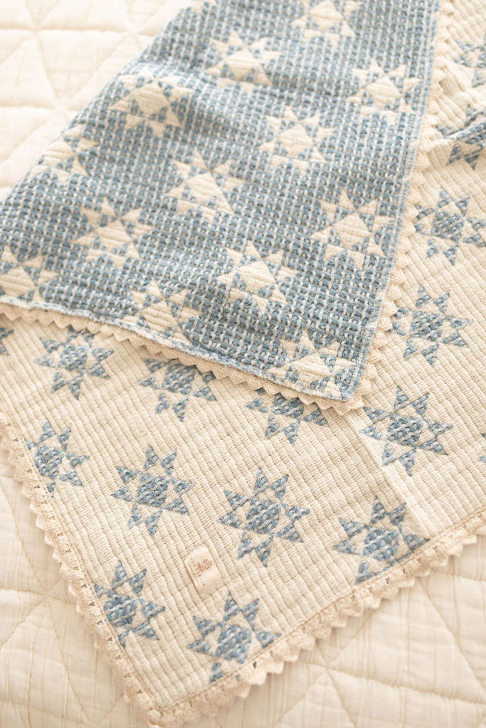The Organic Cotton Baby Star Quilt - Sky features a blue and cream star pattern, textured quilted design, and delicate crochet trim. It’s folded on a cream surface.