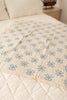 The Organic Cotton Baby Star Quilt - Sky, featuring a cream base with blue star patterns and delicate crochet trim, is draped over a bed with a light quilted coverlet in a softly lit room. Ships in one week.