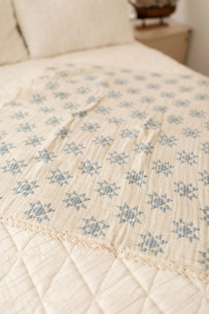 The Organic Cotton Baby Star Quilt - Sky, featuring a cream base with blue star patterns and delicate crochet trim, is draped over a bed with a light quilted coverlet in a softly lit room. Ships in one week.