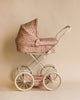 The Konges Sloejd Doll Pram in Cherry Blush features a vintage-style design with quilted fabric and a pleated canopy, set atop four large white wheels that complement its subtly shining metal frame against a plain beige background.