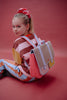 A young girl with a ponytail and pink scrunchie sits on the floor, dressed in a colorful striped outfit and carrying the Sticky Lemon School Bag | Lemon Tree | Gingham Flower + Rose, a pastel bag crafted from waterproof nylon, as she looks over her shoulder against a pink background.
