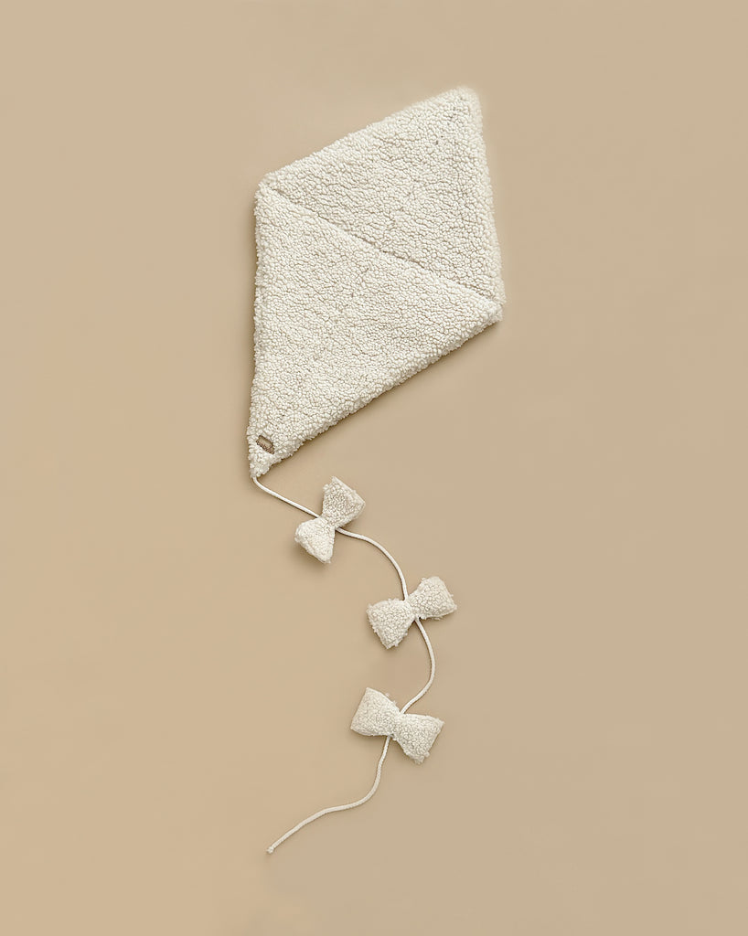 The Boucle Kite Wall Decor - Cream is depicted against a simple beige background. Crafted from Oeko Tex-certified polyester, this triangular, cream-colored fabric kite features a textured, fuzzy surface and a string tail adorned with three bow-shaped fabric ties spaced along it.