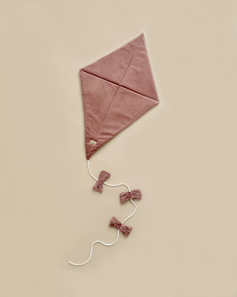 The Velvet Kite Wall Decor in dusty pink, featuring a diamond shape and three velvet bows on its white string, rests elegantly on a beige background—a perfect addition to any child's room decor.