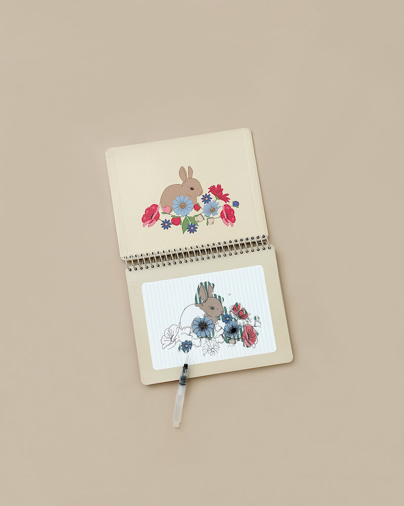 An open "My Magic Water Book - Off White" lies on a beige surface. The top page features an illustration of a brown rabbit surrounded by flowers, while the bottom page shows a partially colored version of the same illustration. A black gel pen rests on the notebook.