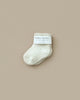 A single fluffy white baby sock from the "Newborn Gift Set - Natural," made with Peruvian cotton yarn and a folded cuff, is displayed on a beige background. The label reads "baby socks" along with other text.