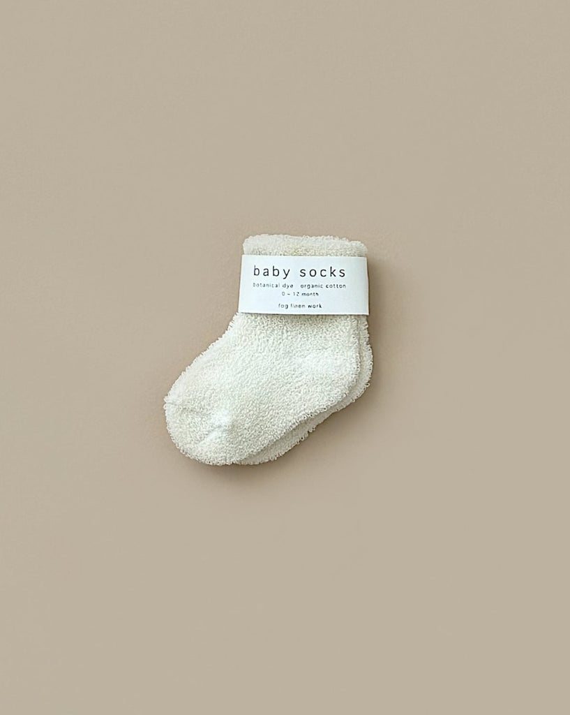 A single fluffy white baby sock from the "Newborn Gift Set - Natural," made with Peruvian cotton yarn and a folded cuff, is displayed on a beige background. The label reads "baby socks" along with other text.
