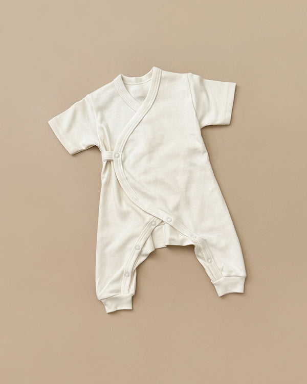 A small white Baby Jumpsuit with short sleeves is laid flat against a beige background. The newborn clothing item features a wrap-style front with snap closures along one side and at the bottom.