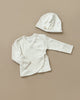 A cream-colored infant outfit, named "Baby Cap & Cardigan," consisting of a long-sleeve wrap shirt with a side button and a matching baby hat, both crafted from soft organic cotton. These cozy items are laid flat on a neutral beige surface.