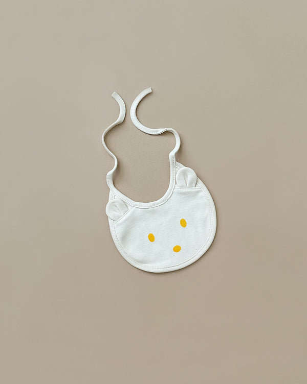 A white organic cotton baby bib, named the Baby Bear Bib, features a simple cat face design with yellow dots for eyes and a nose. It has ties for securing around the neck and is laid flat on a beige surface, making it an ideal newborn gift.