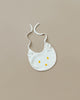 A white organic cotton baby bib, named the Baby Bear Bib, features a simple cat face design with yellow dots for eyes and a nose. It has ties for securing around the neck and is laid flat on a beige surface, making it an ideal newborn gift.