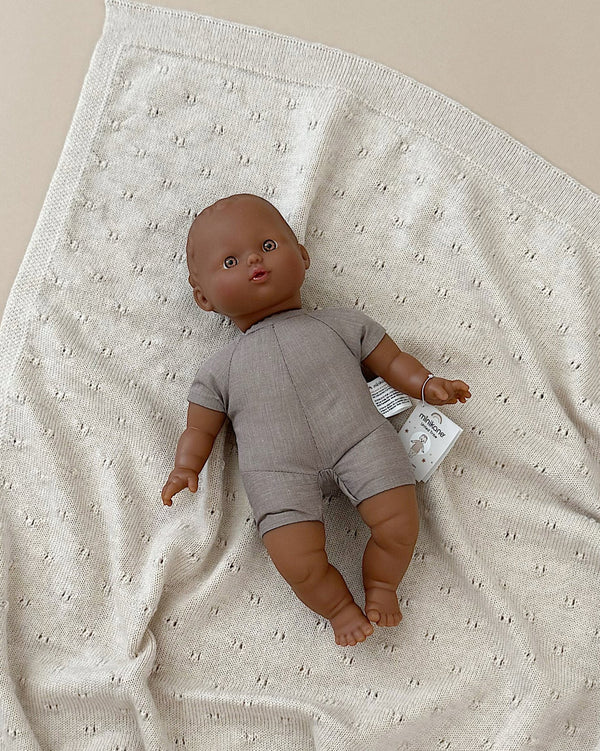 The Minikane Soft Body Doll (11") - Sidonia, crafted from phthalate-free vinyl and featuring a brown complexion with short hair, comes dressed in a beige romper. The doll, which lies on a cream-colored textured blanket with a tag on its left wrist, makes for the perfect bedtime companion against the neutral beige background. It ships in approximately one week.