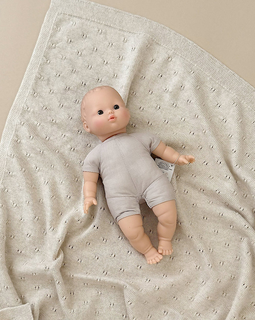 The Minikane Soft Body Doll (11") - Lucien, dressed in a light grey onesie, rests peacefully on a textured cream-colored blanket. Crafted from phthalate-free vinyl, Lucien features a serene expression and lifelike details, with its arms and legs slightly spread out. Ideal for imaginative playtime, the blanket is gently gathered around the doll.