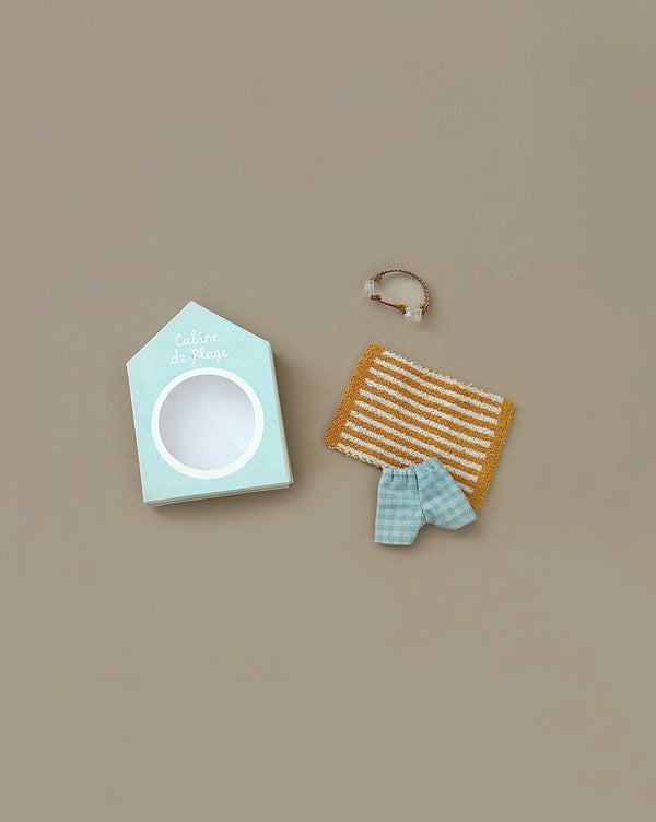 The Maileg Beach Bundle for Big Brother features a light blue beach hut box, a brown and white striped towel, a blue gingham cloth, and a tiny transparent beach bag with gold handles—perfect vacation accessories on a beige background.