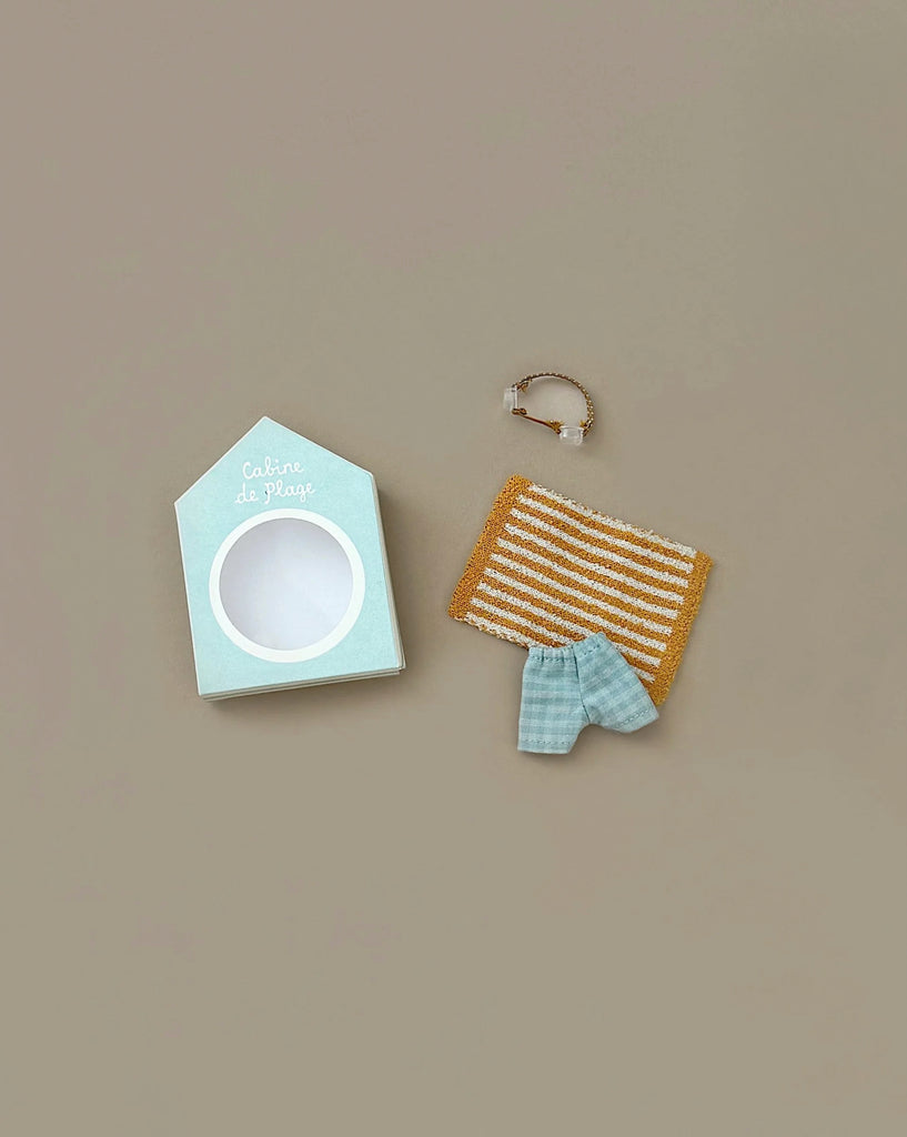 The Maileg Beach Bundle for Big Brother features a light blue beach hut box, a brown and white striped towel, a blue gingham cloth, and a tiny transparent beach bag with gold handles—perfect vacation accessories on a beige background.