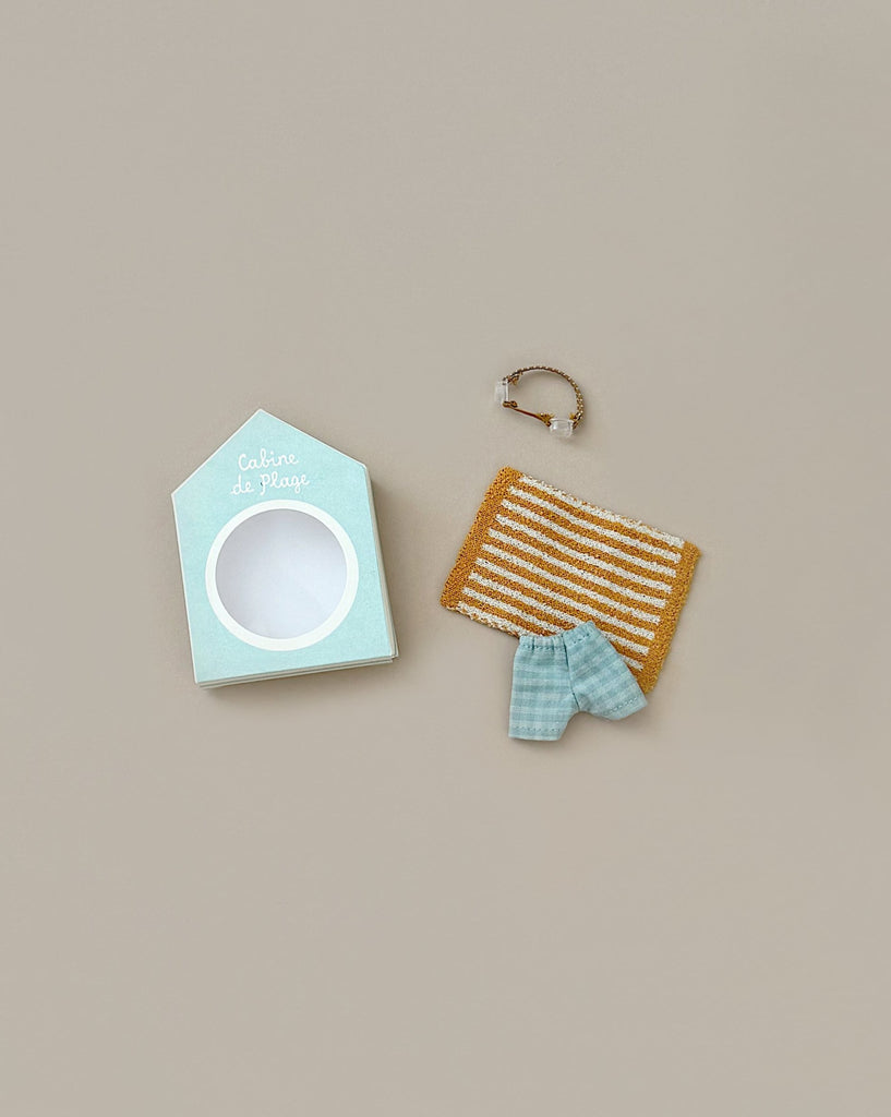 The "Maileg Beach Set for Big Brother," consisting of a cardboard packaging shaped like a beach house labeled "Cabine de Plage," an orange and white striped towel, a small blue and white checkered linen cotton towel, and a bracelet with a pendant, lies on a beige surface.