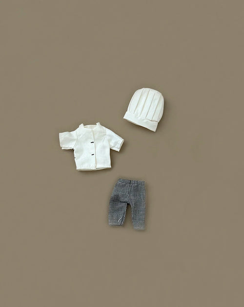 The Maileg Extra Clothing: Chef Clothes For Big Sister And Big Brother Mouse set features a white chef’s hat and jacket with black-and-white checkered pants, displayed on a plain brown background.