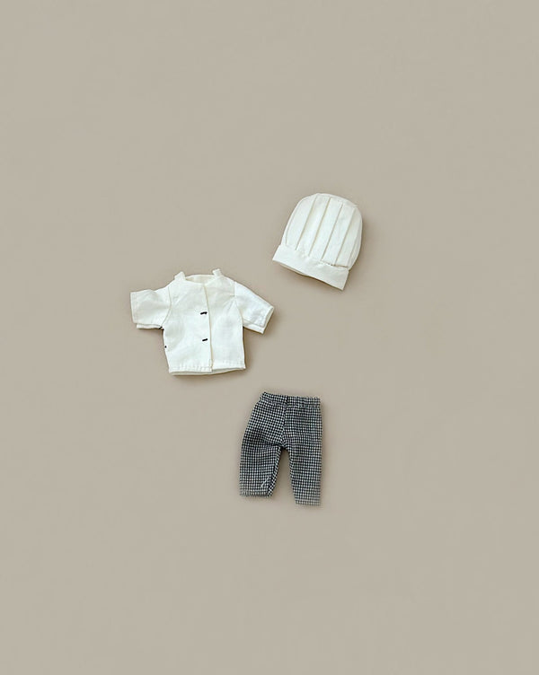 A flat lay of Maileg Extra Clothing: Chef Clothes For Big Sister And Big Brother Mouse on a beige background, featuring miniature chef attire. The set includes a small white double-breasted coat, a pair of tiny checkered pants, and a white quilted hat. Perfectly sized for your big brother mouse or big sister mouse.