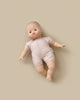 A Minikane Soft Body Doll (11") - Madder, with a bald head and wide, round eyes, is dressed in a simple light beige outfit and lies on a beige surface. Made of phthalate-free vinyl, this doll's slightly spread-out arms and legs make it perfect for imaginative playtime.