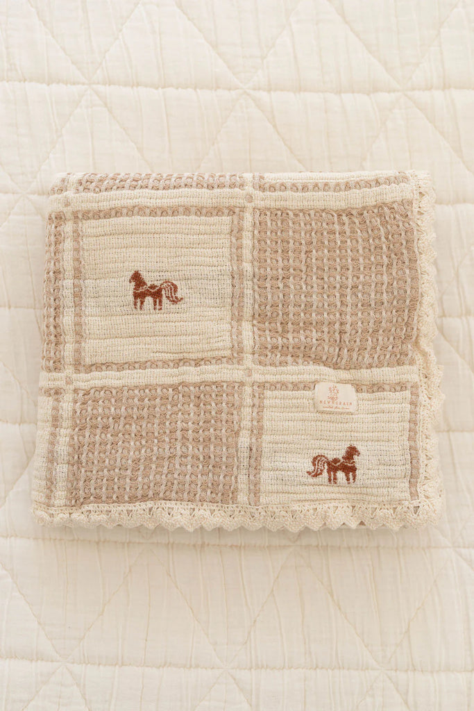 The Organic Cotton Baby Blanket - Pony, featuring a beige textured design with embroidered pony and crochet trim, rests folded on a quilted white surface.