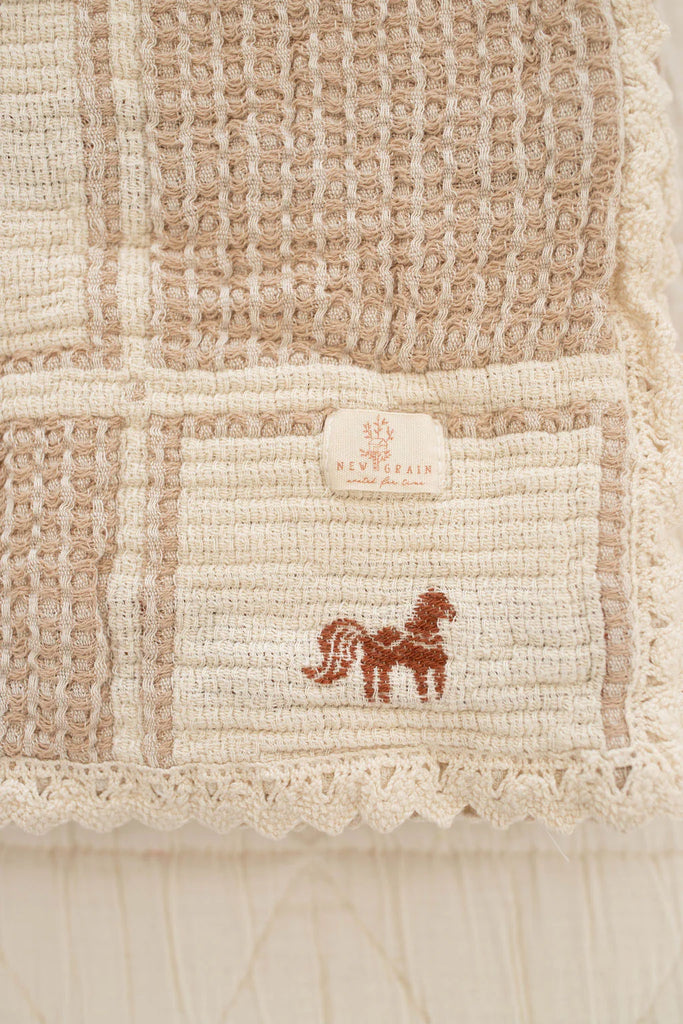 The Organic Cotton Baby Blanket - Pony features a textured beige and cream woven fabric, a small embroidered pony in one corner, a "NEWBRAIN" fabric label above it, and delicate crochet trim for an elegant finish.