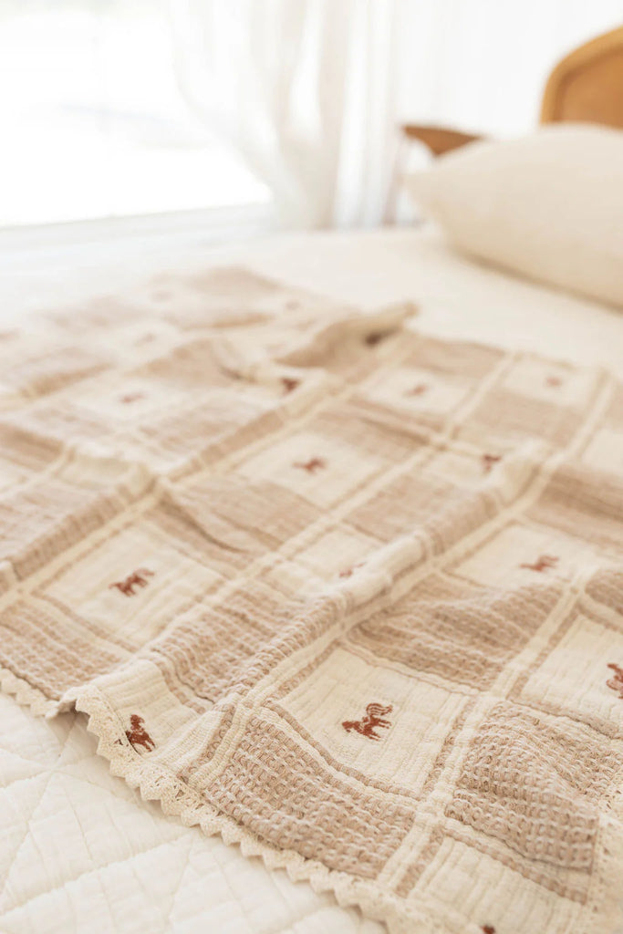 The Organic Cotton Baby Blanket - Pony, featuring beige and cream patchwork with an embroidered pony and crochet trim, is displayed on a white quilted bed in a softly lit bedroom.