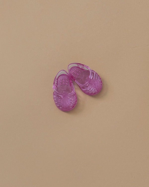 A pair of glitter pink Minikane Beach Sandals, ideal for minikane dolls, are set against a beige background. These delightful sandals infuse a vibrant touch into the neutral setting while embodying the whimsical spirit of French craftsmanship.