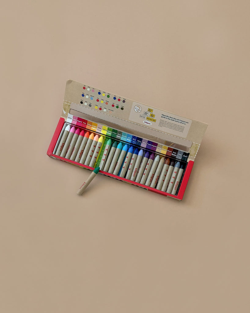 A neatly arranged open box of vibrant Kitpas Crayons - 24 Colors sits on a light beige surface, with one green crayon slightly out of place, creating a serene scene in the background.